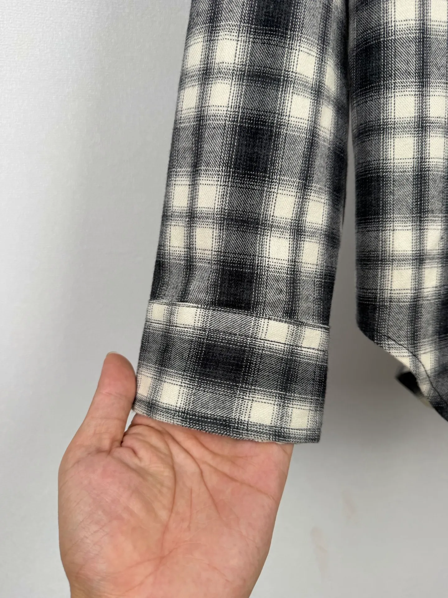 BALENCIAGA/balenciaga Black White plaid pocket embroidered logo shirt jacket The technical difficulties of this plaid shirt focus on fabric finishing, plaid matching and component sewing details. The fabric has been sanded and distressed to create a fine velvet feel on the surface, changing the original luster of the fabric and making the Black and White plaid colors transition softly. At the same time, the drape properties of the fabric itself are retained, taking into account the looseness of the upper body and the silhouette of the body, without appearing soft and shapeless. The biggest test of plaid garments comes from the alignment of the pieces. The front and rear body, large and small sleeves, and chest pockets are all independent pieces. In the cutting stage, the horizontal and vertical axes of the plaid need to be aligned, and the external chest bag is cut separately. The plaid on the bag surface needs to form a visual coordination with the main body of the garment. Cutting and typesetting will produce additional fabric loss, and there are high requirements for the cutting accuracy of the cutting bed typesetting and workers. The collar adopts a double-layer combined structure, and the collar surface and collar seat are double-stitched with even and regular stitching spacing to ensure the lapel's long-lasting shape and resistance to warping and deformation after repeated bending. The placket is sewn with regular topstitching and paired with Black matte buttons. The button points need to be aligned with the checkered lines to avoid damaging the overall visual order. The splicing of the cuffs also takes into account the grain direction, and the overlocking process reinforces the seams to improve the durability of the garment. After the entire garment is sewn, it also undergoes a garment washing process to further strengthen the Retro texture of the fabric, unify the tone and texture of the entire garment, weaken the harshness of industrial production, and finally present a relaxed Retro garment effect. Size: 46 48 50 52